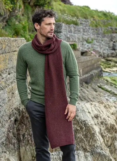 Man wearing a burgundy wool cashmere tweed scarf draped over a green sweater, standing on a rocky coastal path in Ireland.